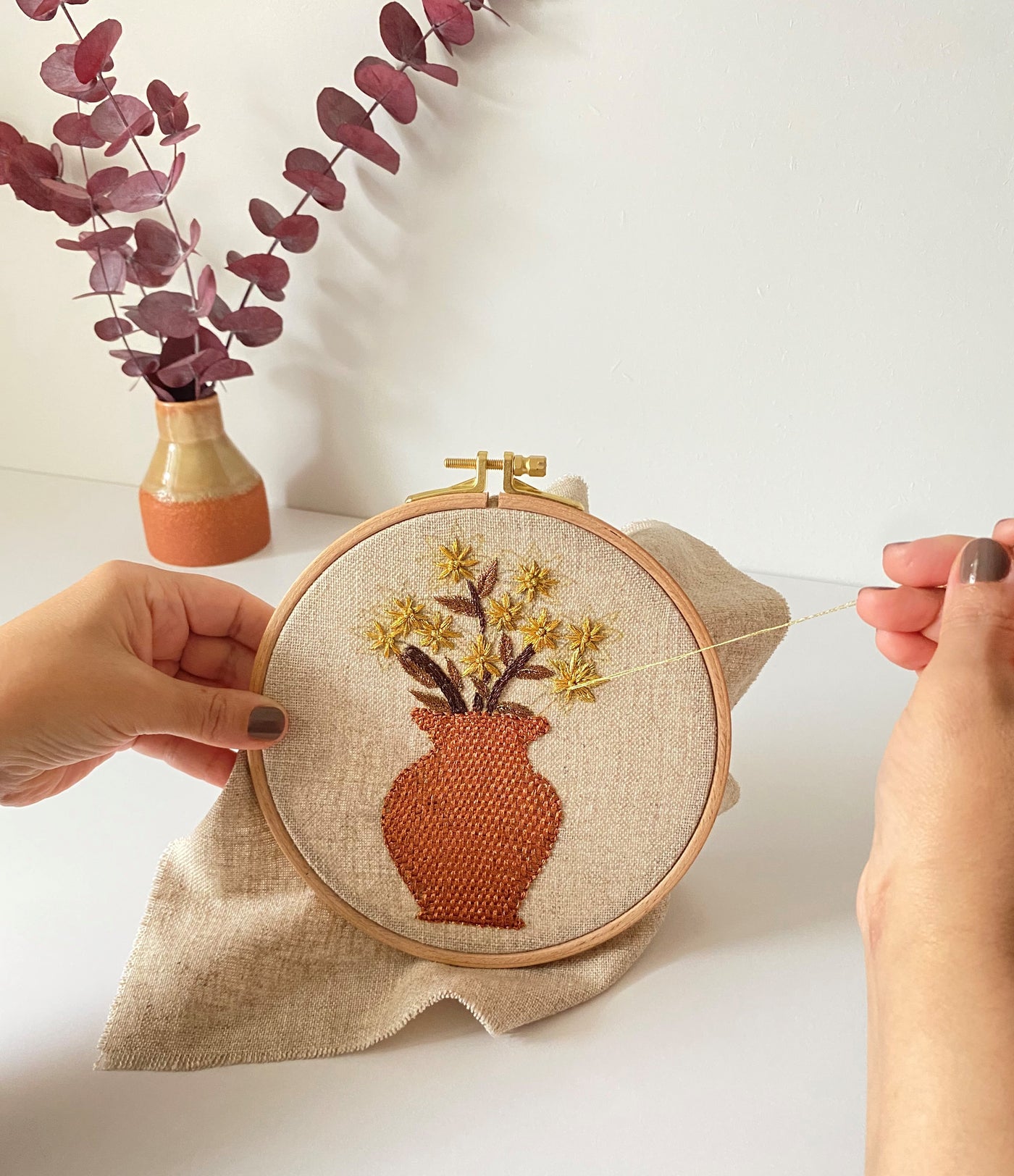 Hands holding the terracotta vase hoop above a linen pouch, beside dried eucalyptus in a vase