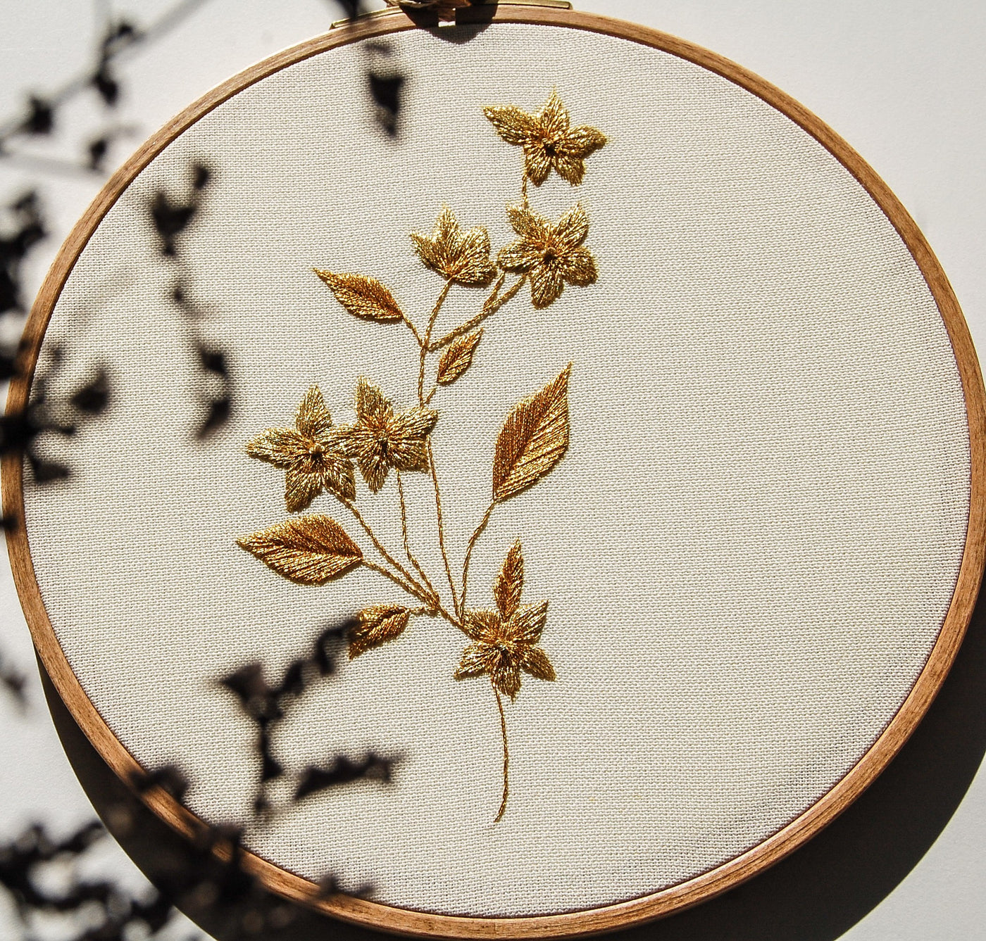 Gilded blossom branch worked in gold metallic thread in a wooden hoop, with leaf shadows falling across the linen