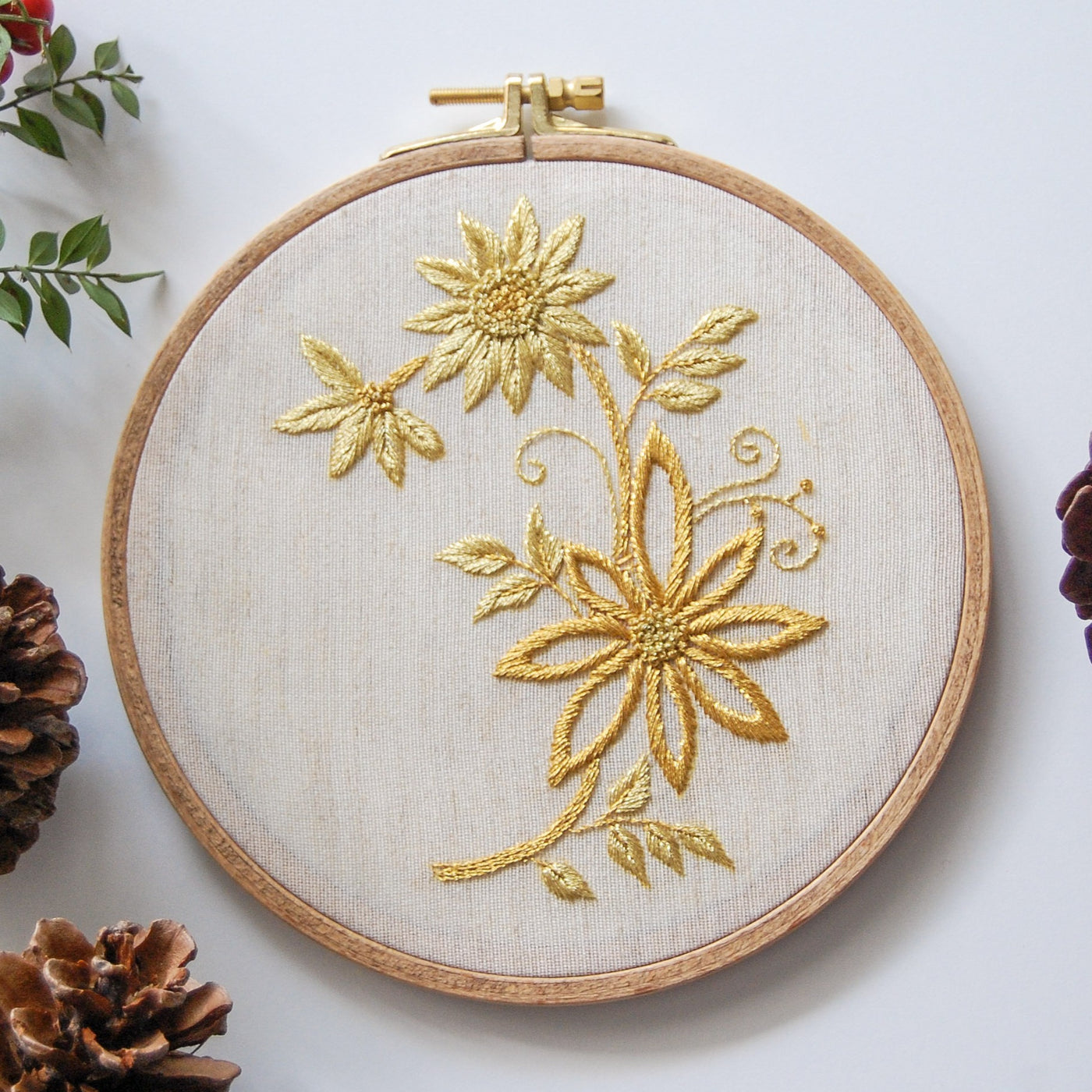 Golden daisies and curling tendrils worked in metallic thread in a wooden hoop, styled with pine cones and greenery