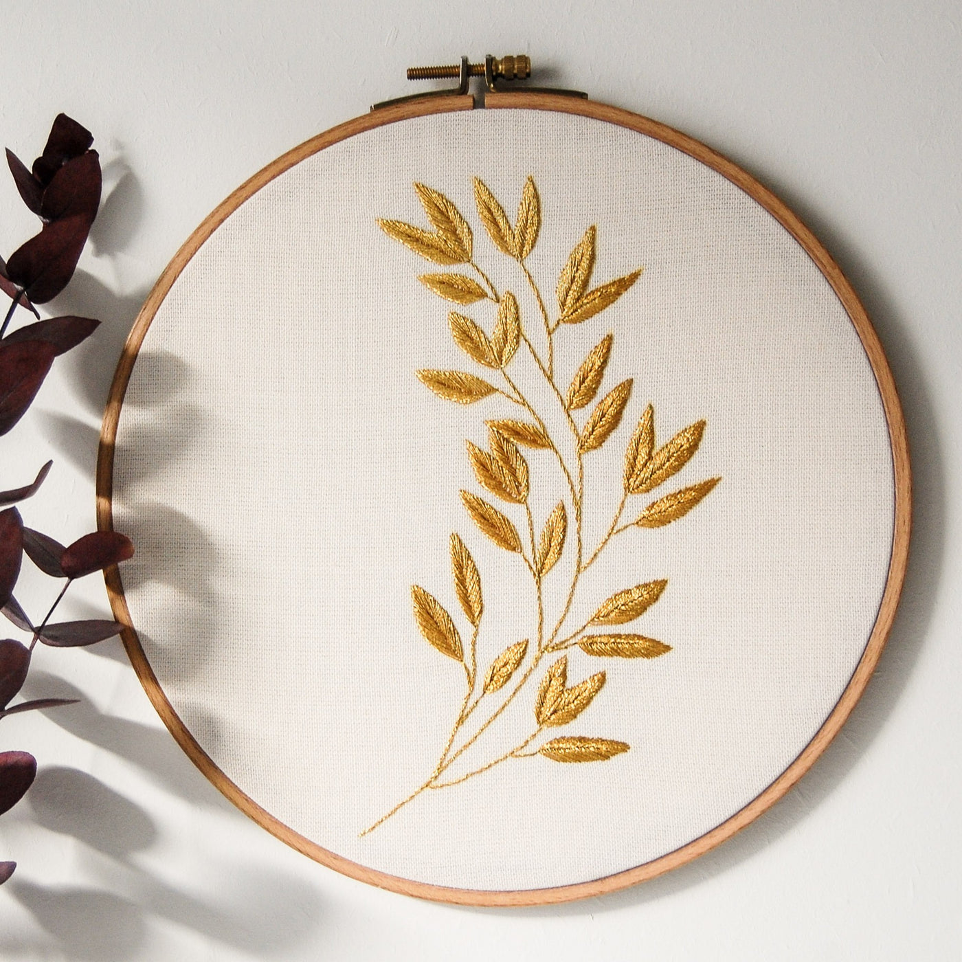 A single golden laurel sprig embroidered in metallic thread in a wooden hoop on ivory linen