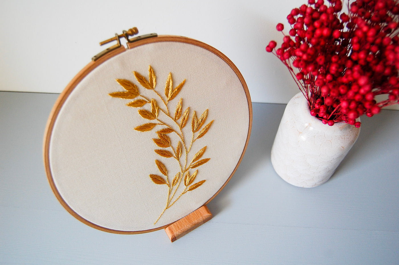 The golden laurel sprig hoop on a stand beside red berries in a white vase
