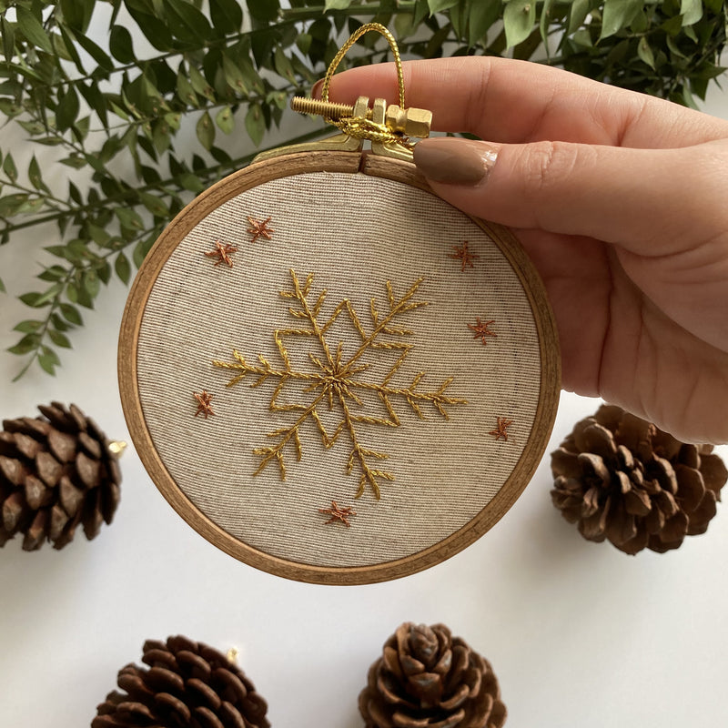 A hand holding a small hoop with a golden snowflake and scattered stars, with pine cones and greenery