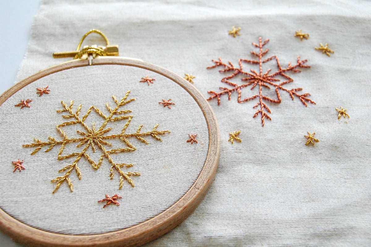 The golden snowflake hoop on white cloth beside the same snowflake stitched in a rust shade