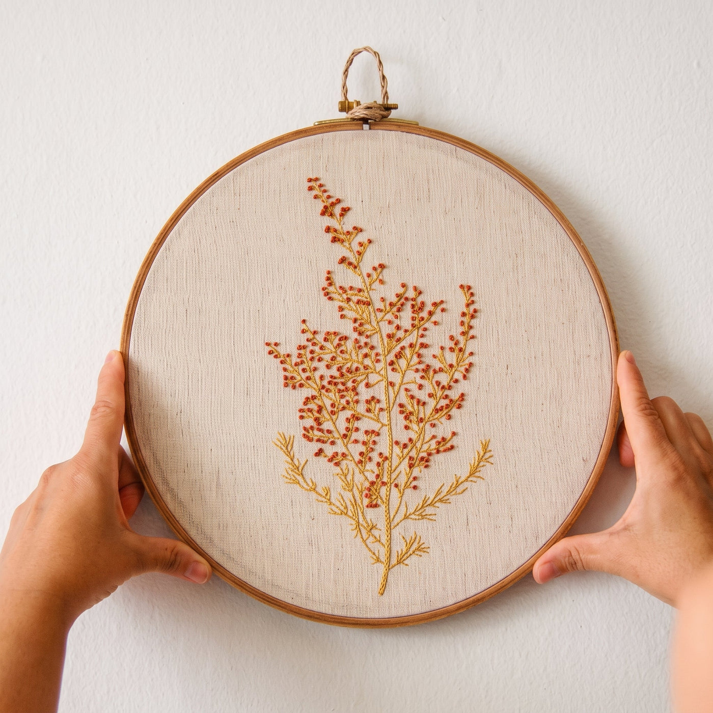 Hands holding a hoop with a tall wildflower spray embroidered in orange and gold thread
