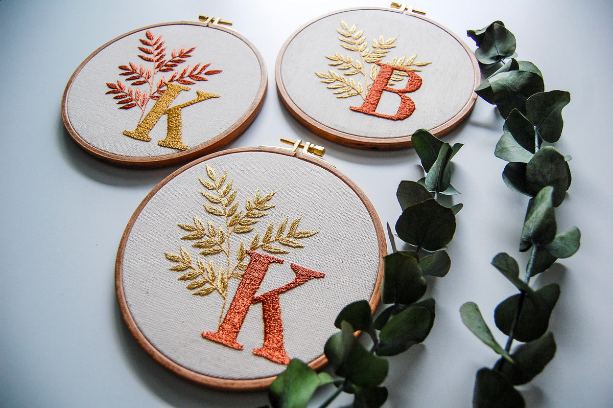 Three monogram hand embroidery hoops with leafy sprigs, beside eucalyptus stems