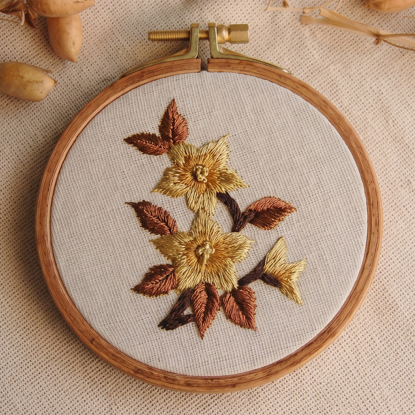 Two golden star flowers with amber and brown leaves embroidered on natural linen in a wooden hoop