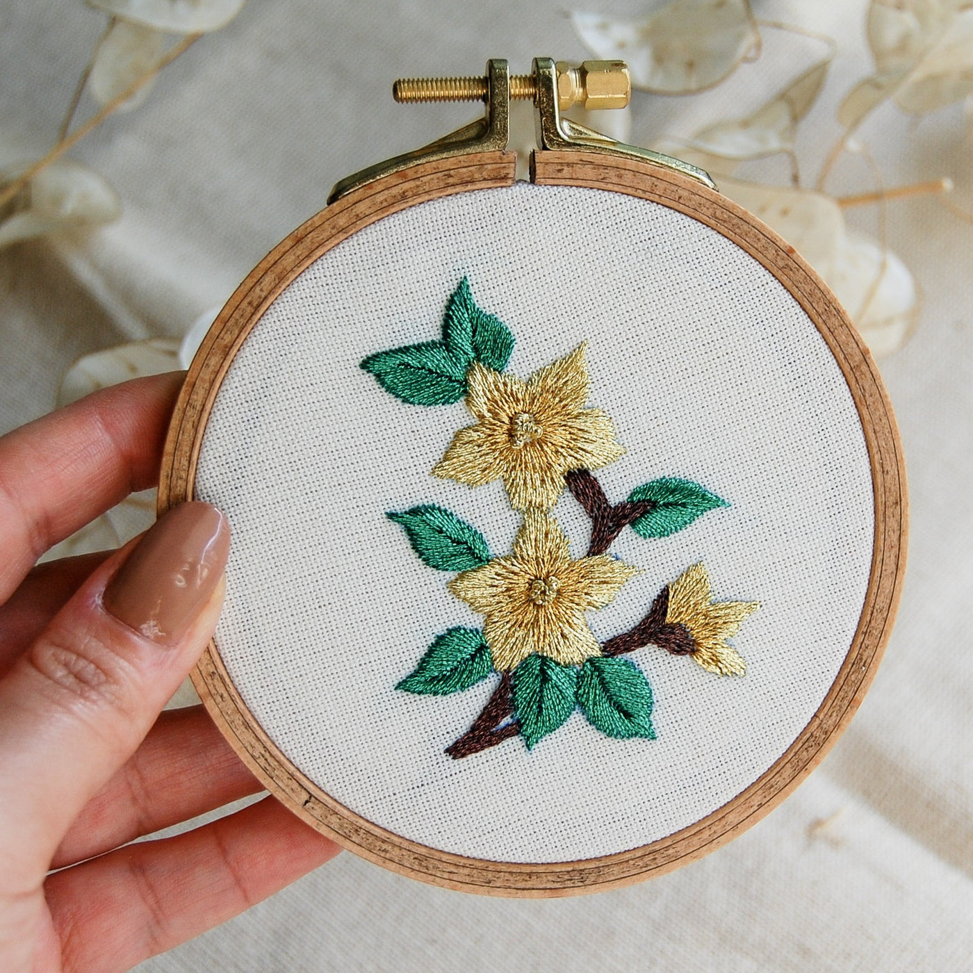 A hand holding a hoop with golden star flowers and jade green leaves on natural linen