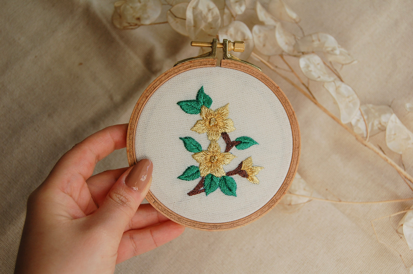 A hand holding the jade mini golden blooms hoop against soft cream fabric
