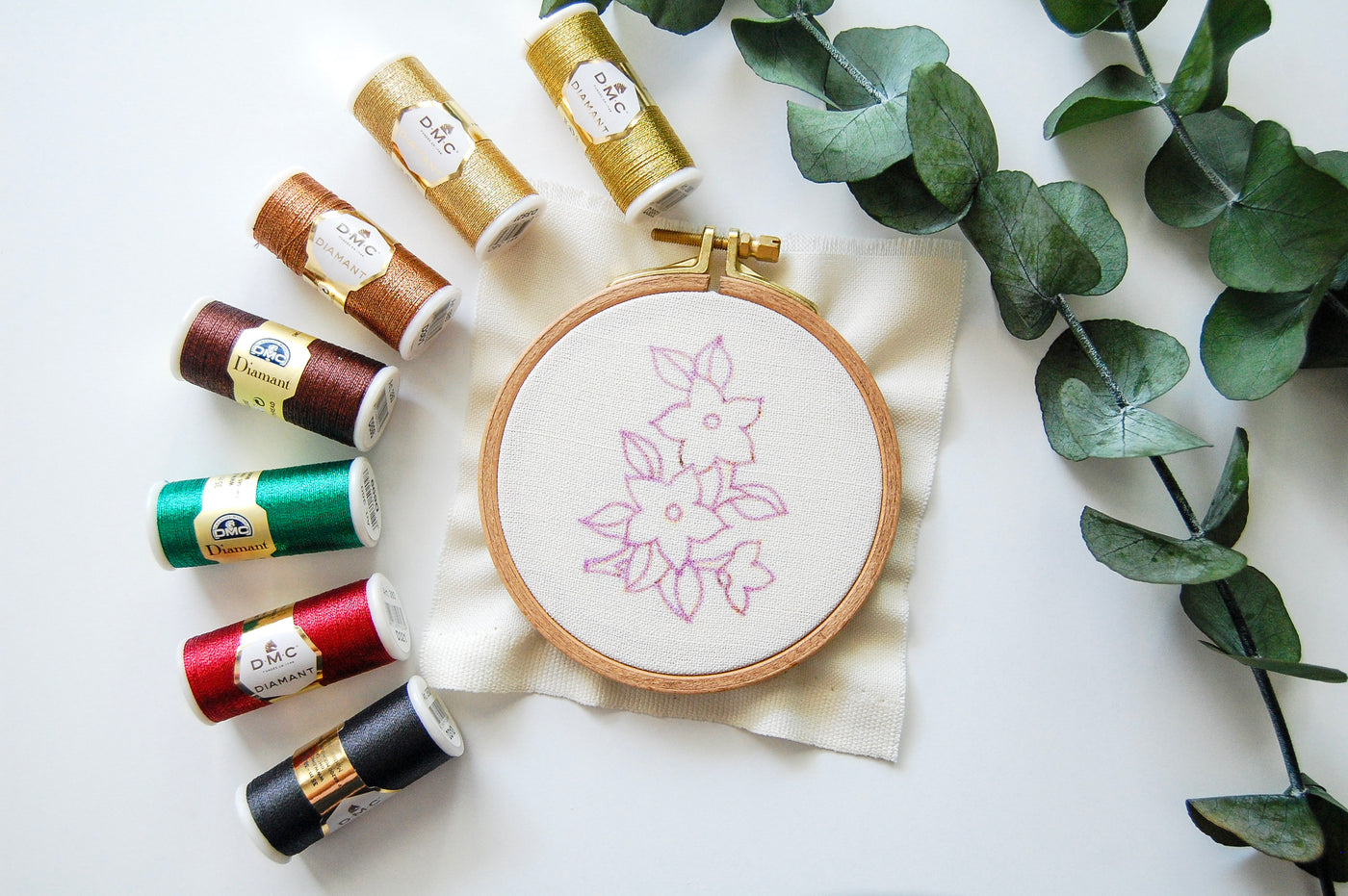 The mini golden blooms pattern on fabric in a hoop, with spools of thread and eucalyptus